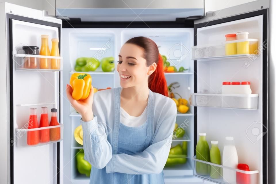 Jovem mulher com pimentão perto de geladeira aberta