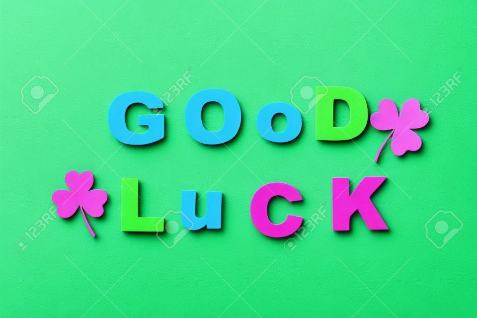 Phrase GOOD LUCK made of color letters and clover leaves on green background, flat lay