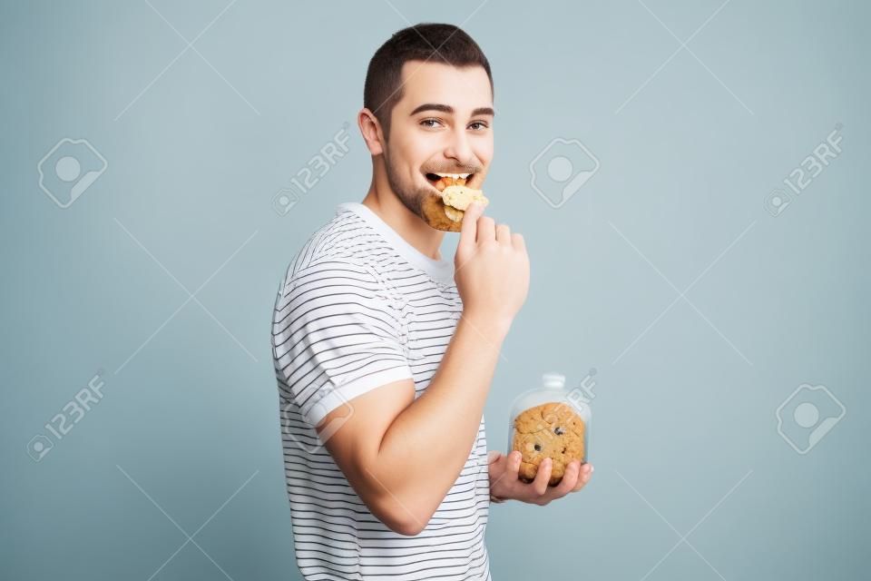 Jeune homme mangeant un biscuit et tenant une jarre à biscuits isolé sur fond blanc