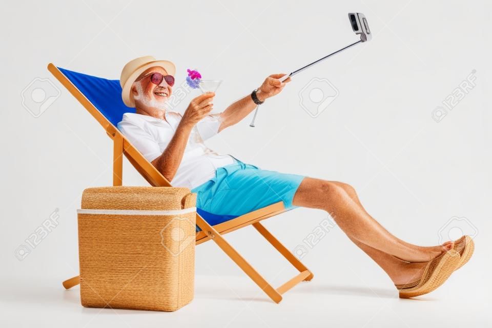 Happy elderly tourist with a cocktail sitting in a sun lounger and taking a selfie isolated on white background