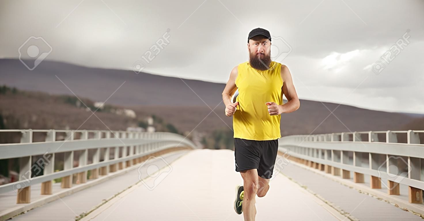Homme barbu avec une casquette faisant du jogging sur un pont