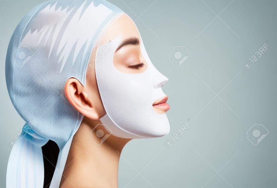 Femme application d'un masque facial feuille pour le traitement de son visage. Gros plan, vue de côté.