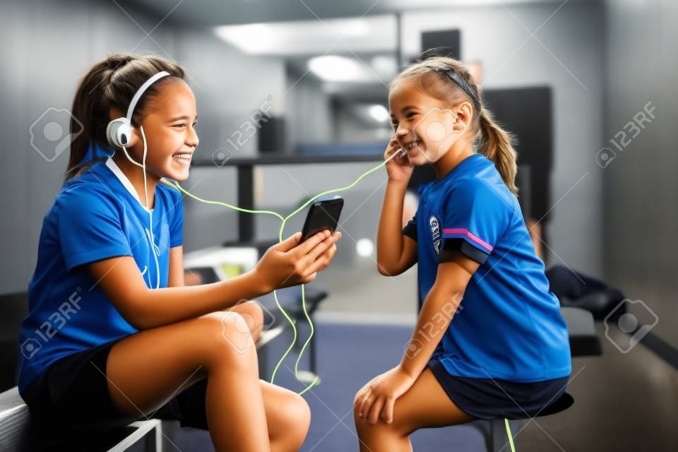 라커룸에서 전화로 훈련하기 전에 두 명의 웃는 여자 축구 선수가 음악을 듣습니다.