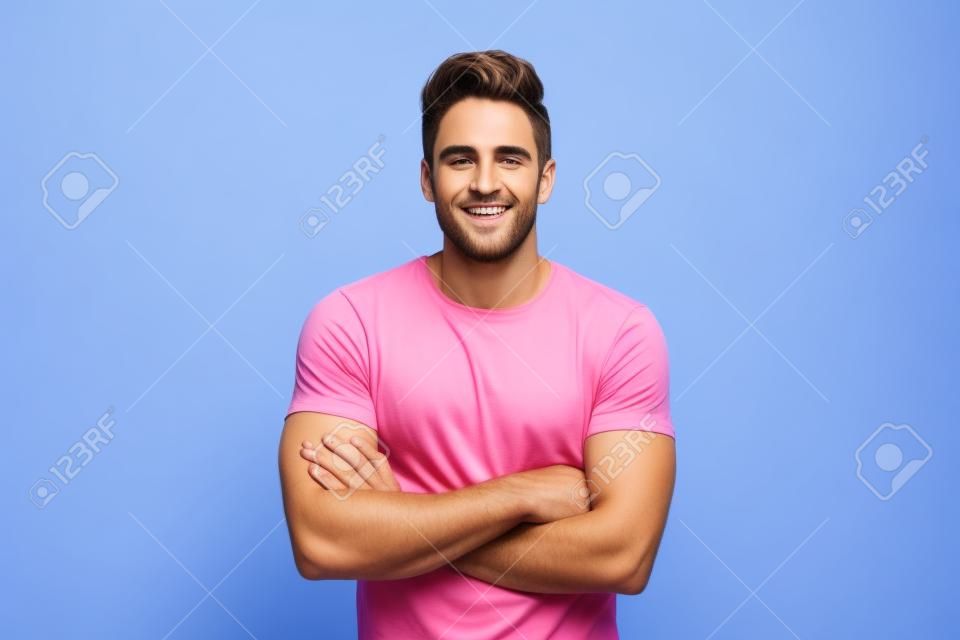 Apuesto joven en camisa rosa sobre fondo azul aislado manteniendo los brazos cruzados en posición frontal