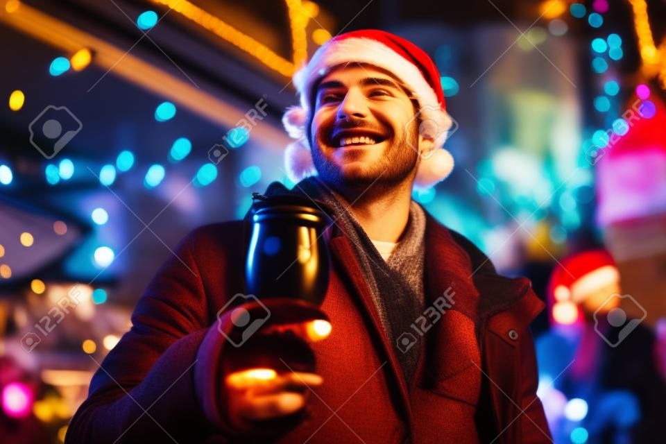 Happiness. Smiling young man holding cup of mulled wine on christmas market, street fair, outdoor. Holidays, emotions, hobbies, leisure activities. Night sky with lights