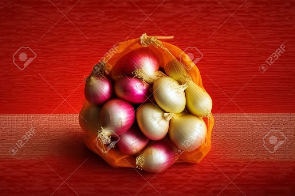 5 kg farm onions in a red pp mesh bag. Polypropylene net sack with 11 lb of organic onions on a brown floor indoors. Buying fresh vegetables in bulk. Front view.