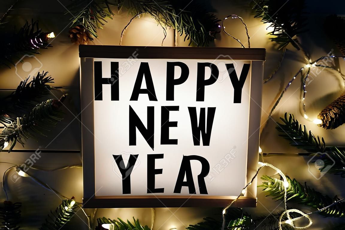 Happy New Year text displayed on a light box on the table with Christmas lights and fir branches. Night. New Year concept.