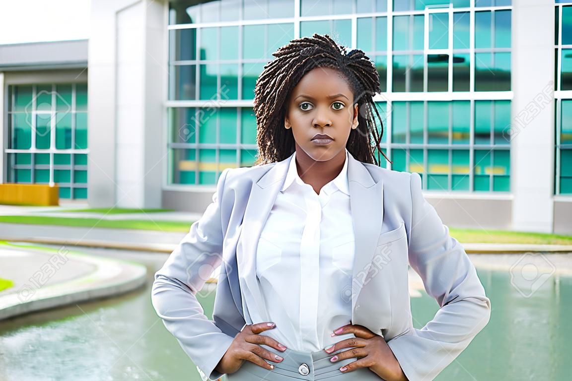 Confident determined businesswoman posing outside. Young black woman wearing formal suit, standing near office building and looking at camera. Confident professional concept