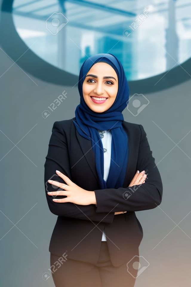 Happy successful Muslim businesswoman posing outside. Beautiful young business woman in black hijab and office suit standing for camera with arms folded and smiling. Muslim business lady concept