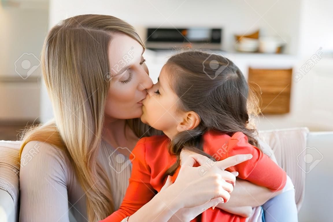 Mom sitting with her little girl on couch, holding kid in arms, kissing and embracing her. Mother enjoying leisure time with daughter at home. Motherhood or family concept