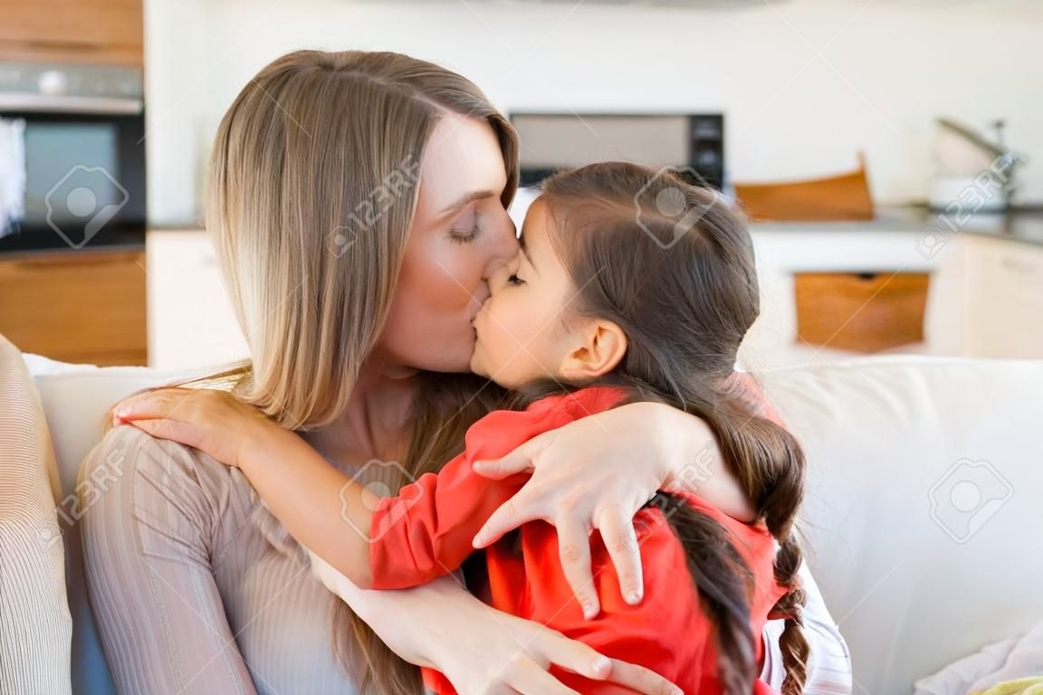 Mom sitting with her little girl on couch, holding kid in arms, kissing and embracing her. Mother enjoying leisure time with daughter at home. Motherhood or family concept
