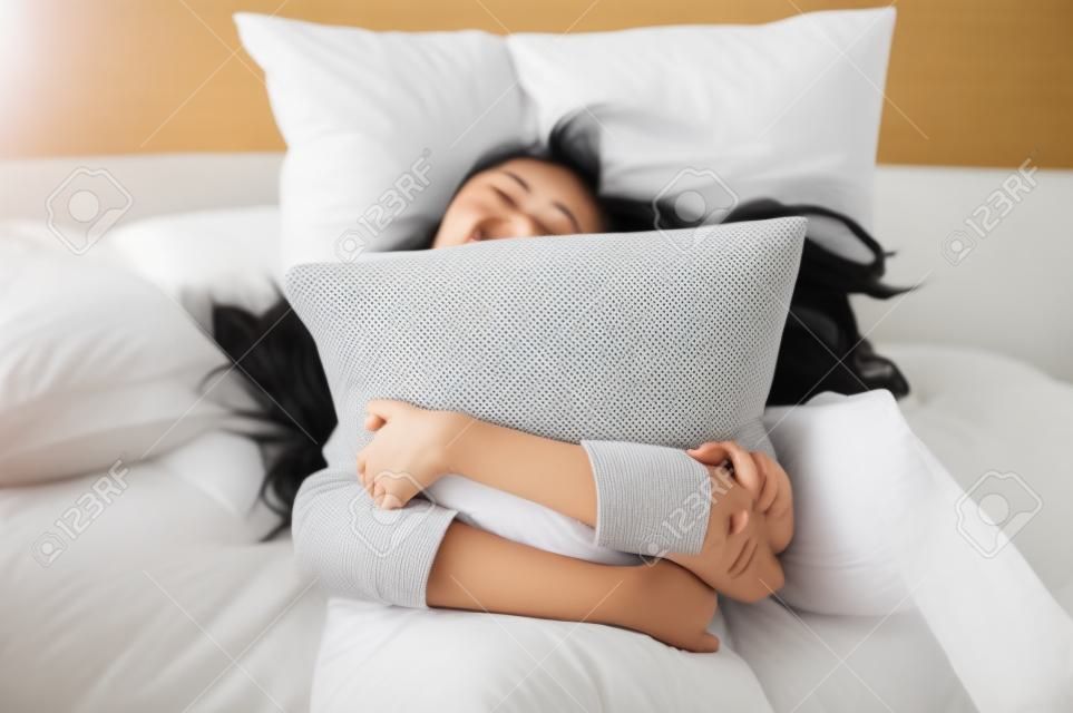 幸せなアジアの若い女性は朝のベッドで枕を抱きしめます