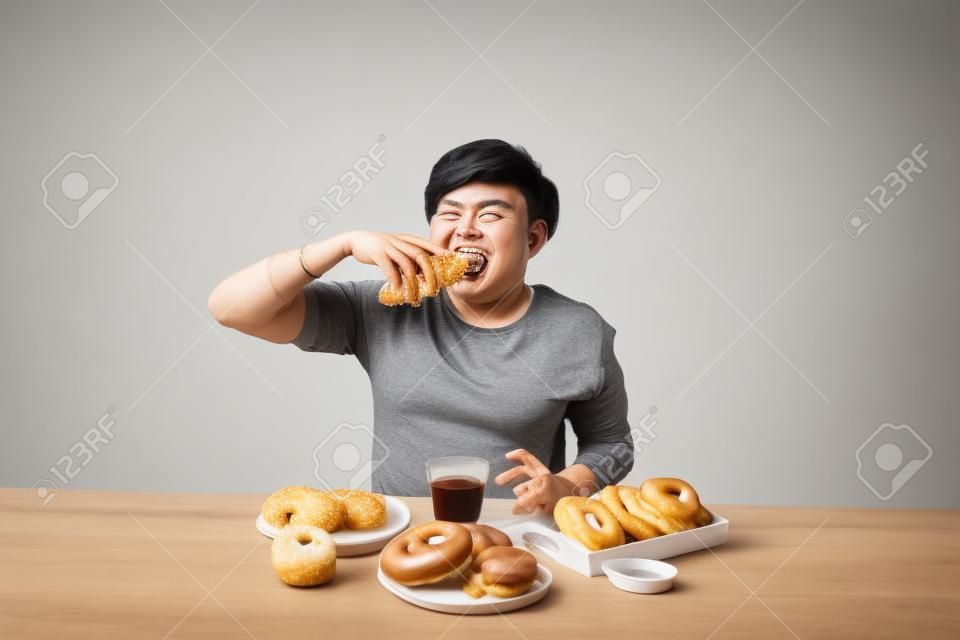 Retrato do homem asiático que senta-se e que petisca donuts isolado sobre o fundo branco.