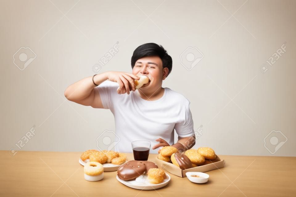 Retrato do homem asiático que senta-se e que petisca donuts isolado sobre o fundo branco.