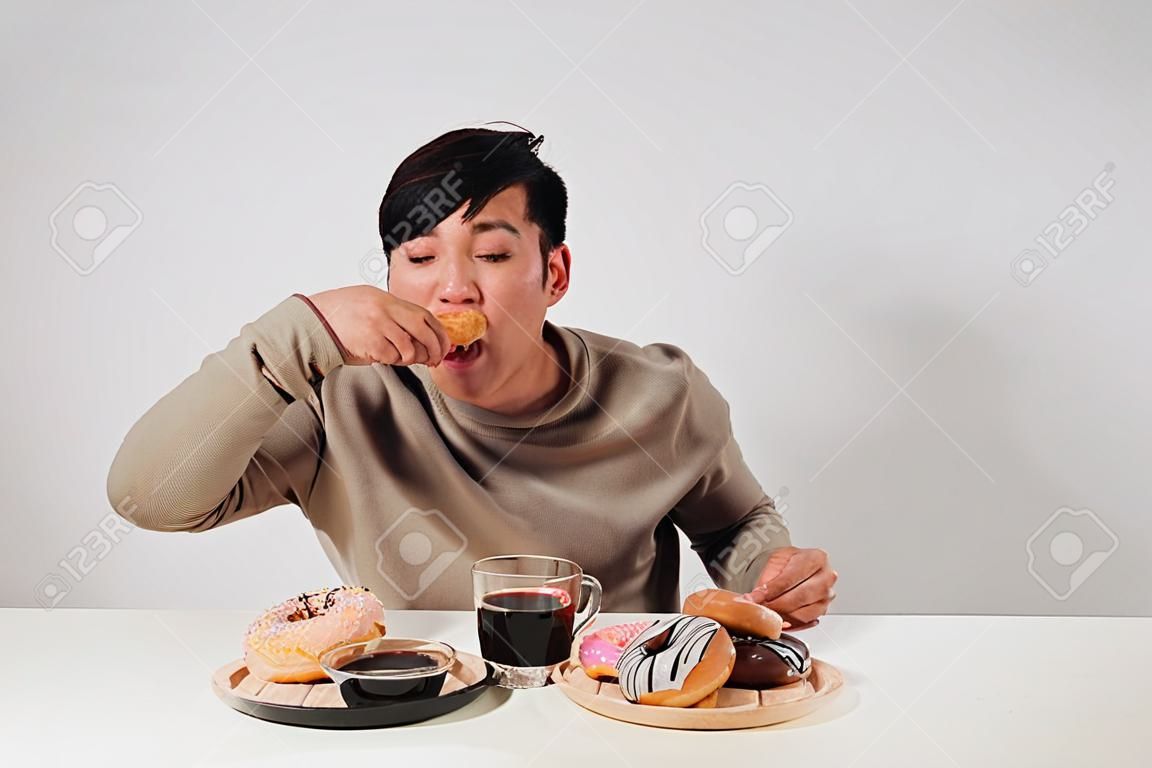 Porträt des asiatischen Mannes, der Donuts lokalisiert über weißem Hintergrund sitzt und isst. Diätkonzept für fette Männer