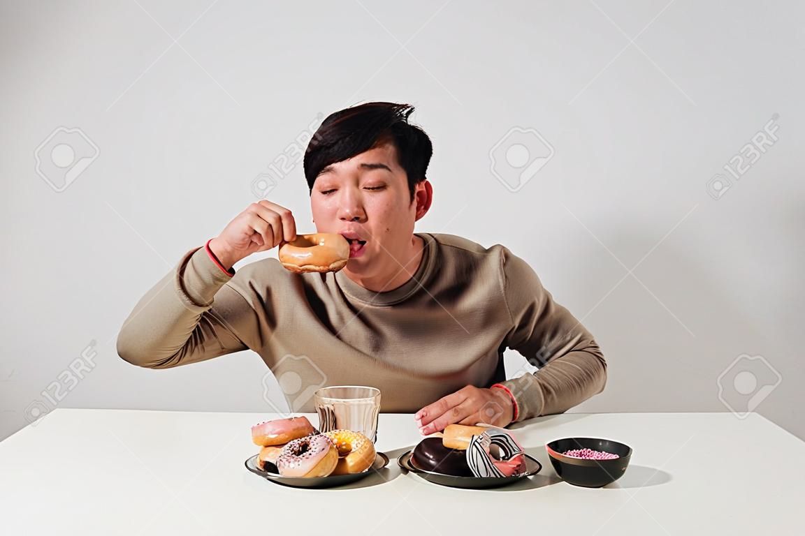 白い背景の上に孤立した座ってスナックドーナツを食べるアジア人男性の肖像画。太った男ダイエットの概念
