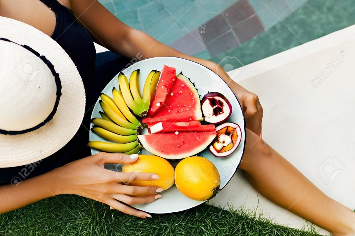 Young unrecognizable woman by the pool with a plate of tropical fruits: watermelon, pineapple, bananas, mangosteen, passion fruit, mango and dragon fruit.