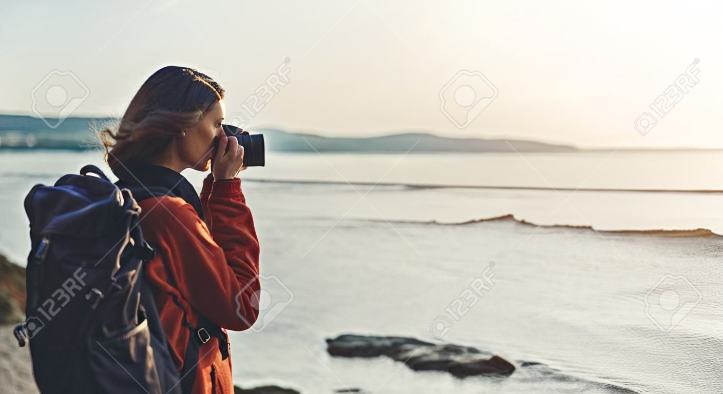 Hipster hiker tourist with backpack taking photo of amazing seascape sunset on camera on background blue sea, photographer enjoying ocean horizon, blurred panoramic sunrise blank mockup, traveler relax holiday concept, sunlight view in trip vacation northern spain 