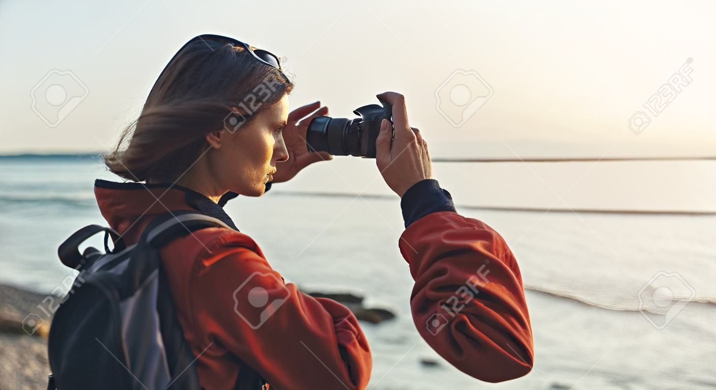 Hipster hiker tourist with backpack taking photo of amazing seascape sunset on camera on background blue sea, photographer enjoying ocean horizon, blurred panoramic sunrise blank mockup, traveler relax holiday concept, sunlight view in trip vacation northern spain 