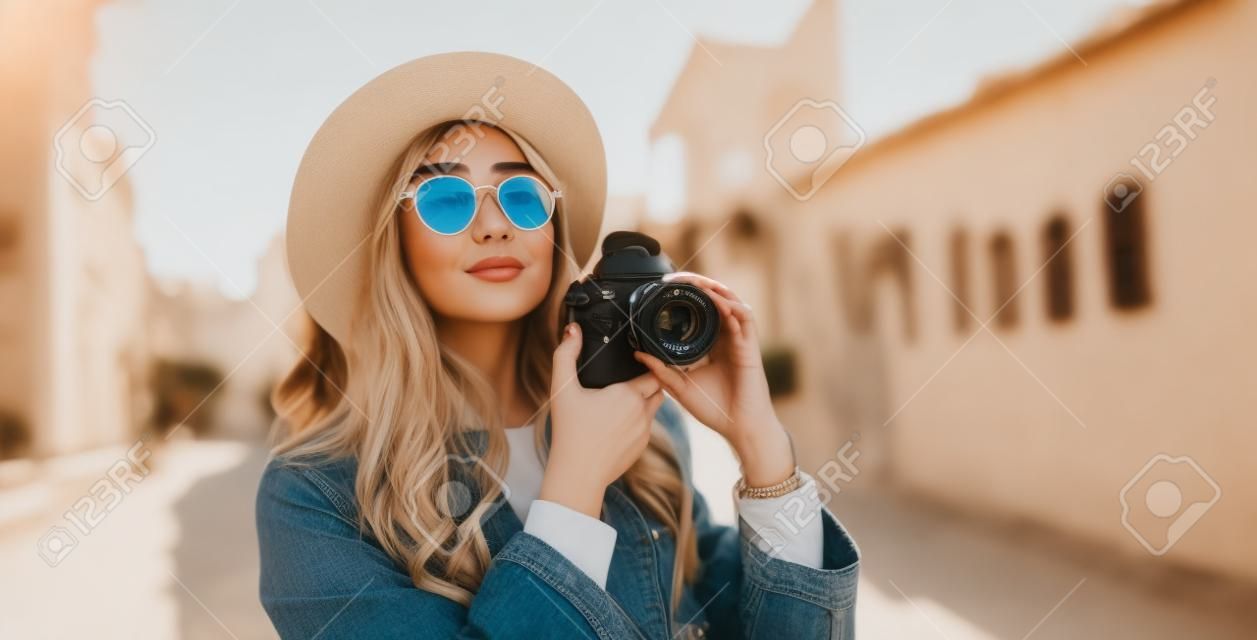 Hobby photographer concept. Outdoor lifestyle portrait of pretty woman in sun city in Europe with camera travel photo of photographer in glasses and hat take photo copy space mockup