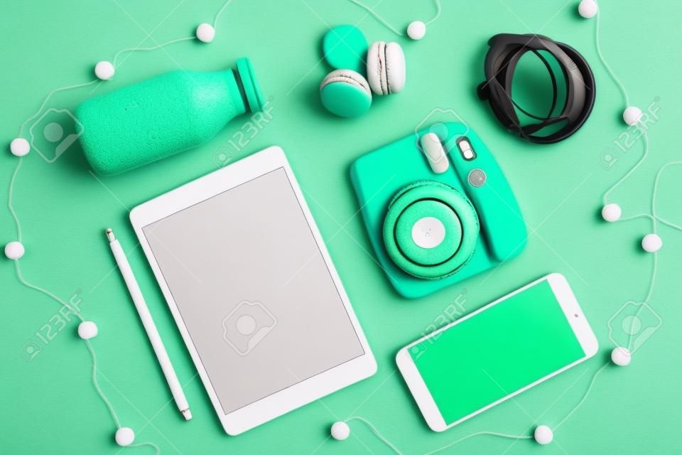 Flat lay tablet, phone, analog camera and headphones, bottle and macaroons on green background