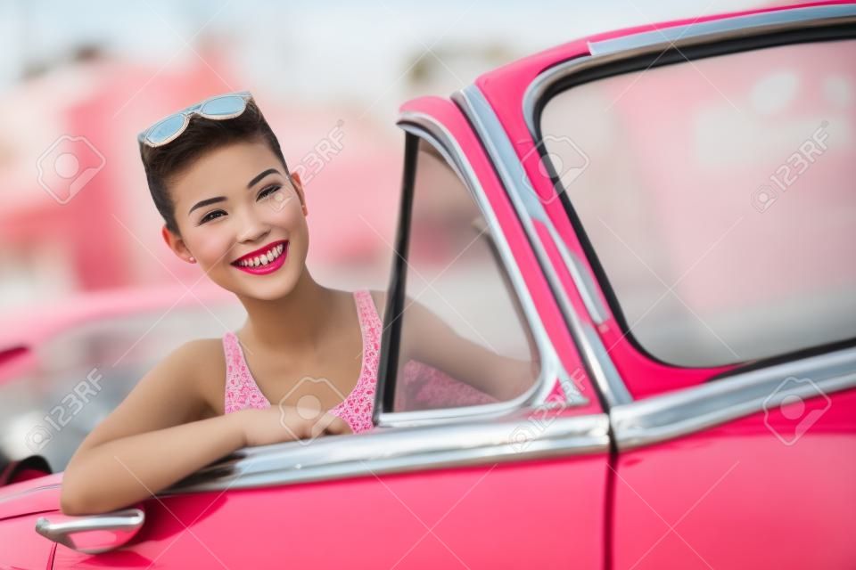 Woman Driving Oldtimer. Retro-Stil Bild der glücklich lächelnde junge Frau in rosa alten Auto, das auf Road Trip an sonnigen Frühlings-oder Sommertag. Schöne multirassischen Kaukasier / chinesische asiatische Lifestyle-Modell im Freien. Szenenfoto aus Havanna, Kuba.