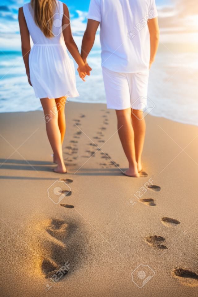 Pareja de la mano caminando romántica en la playa de vacaciones vacaciones viaje dejando huellas en la arena. Primer plano de los pies y la arena dorada de espacio de la copia. Pareja joven con pantalones cortos blancos.
