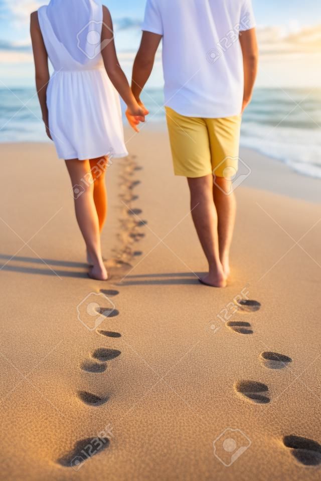 Trzymając się za ręce para spaceru romantycznych wakacji na plaży na wakacje pozostawiając ślady na piasku. Zbliżenie stóp i złoty piasek na przestrzeni kopii. Młoda para ma na sobie białe szorty.