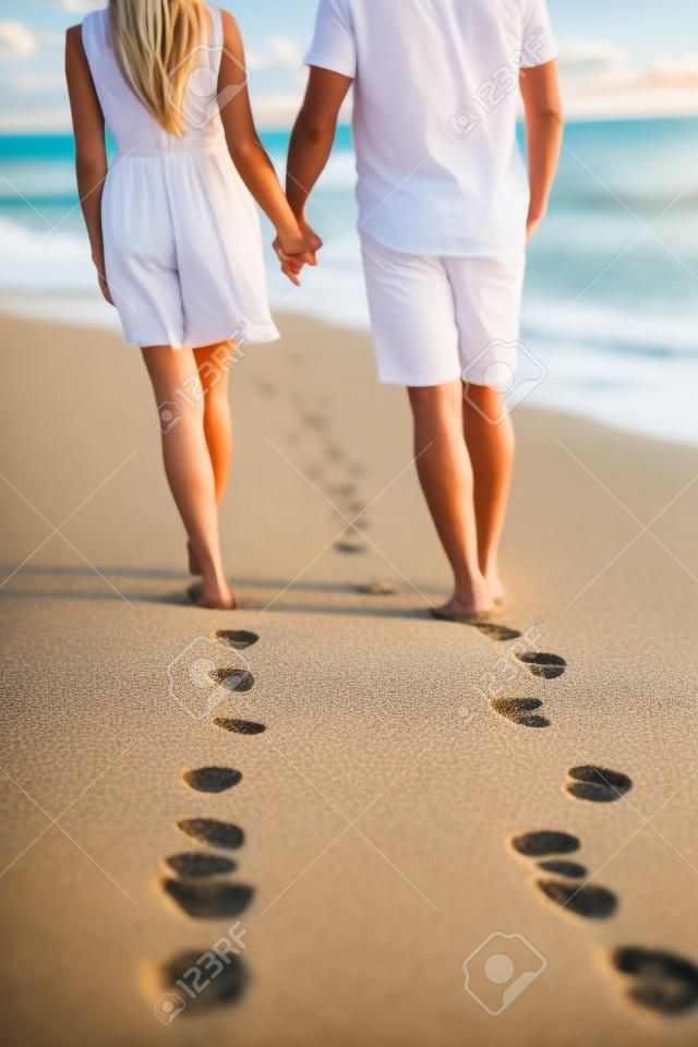 Trzymając się za ręce para spaceru romantycznych wakacji na plaży na wakacje pozostawiając ślady na piasku. Zbliżenie stóp i złoty piasek na przestrzeni kopii. Młoda para ma na sobie białe szorty.