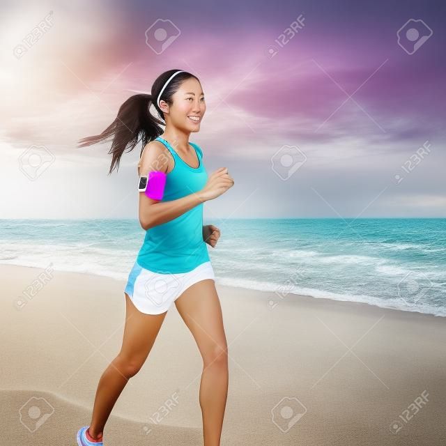 Donna corrente che pareggia sulla spiaggia che ascolta la musica in trasduttori auricolare dal braccialetto di smartphone del riproduttore mp3 del telefono cellulare, addestramento femminile del corridore per la maratona sulla bella spiaggia. Donna asiatica razza mista