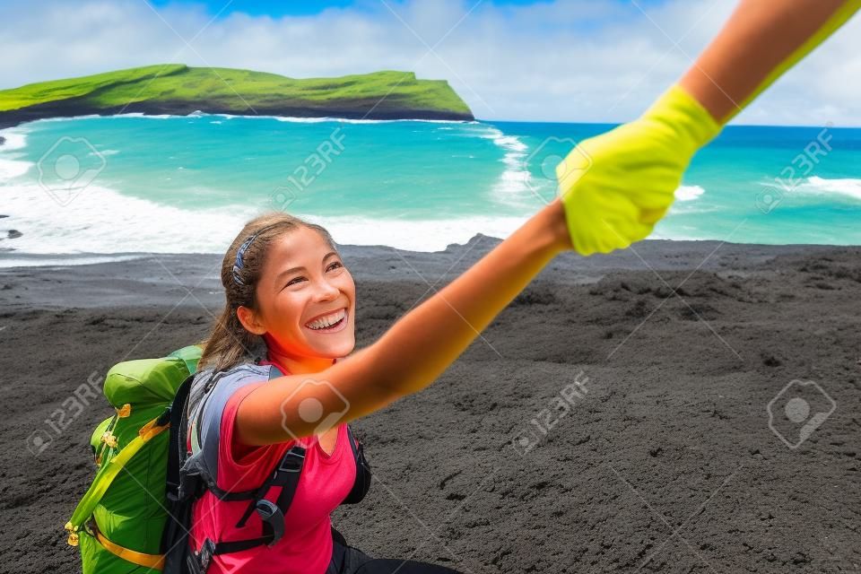 Mano amiga - excursionista mujer recibiendo ayuda en caminata sonriendo feliz superando obstáculo. Mochileros turísticos caminando en la playa de arena verde, Papakolea en Big Island, Hawaii, Estados Unidos. Pareja joven viajando.