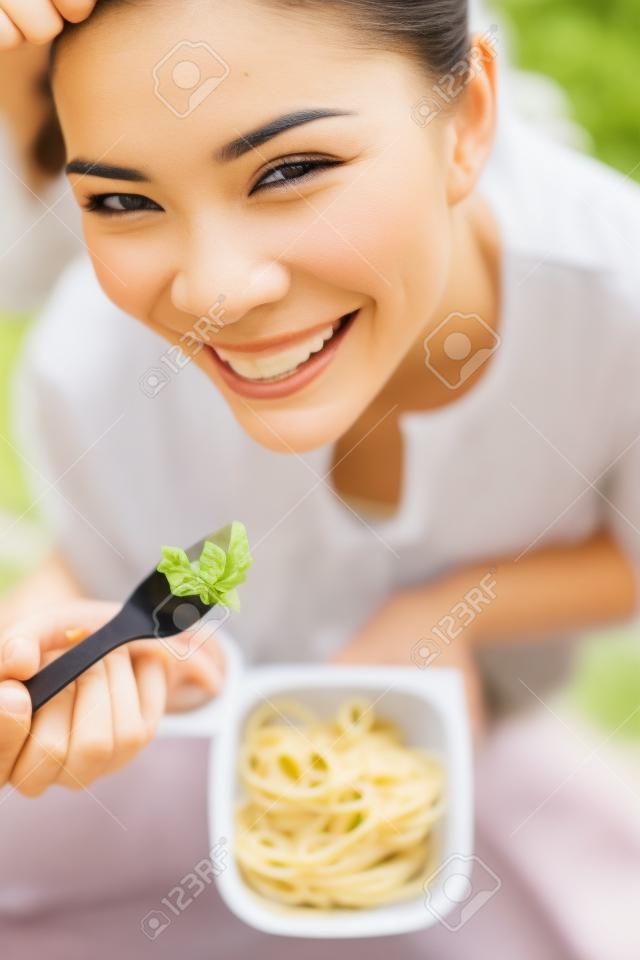 Mujer de comer gluten ensalada de pasta gratis. Retrato de niña comiendo comida para llevar en el parque al aire libre. Modelo femenino birracial asiática del Cáucaso en sus veinte años sonriendo feliz mirando a la cámara.