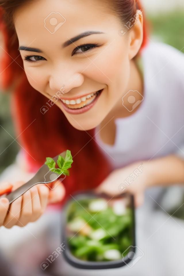 Mujer de comer gluten ensalada de pasta gratis. Retrato de niña comiendo comida para llevar en el parque al aire libre. Modelo femenino birracial asiática del Cáucaso en sus veinte años sonriendo feliz mirando a la cámara.