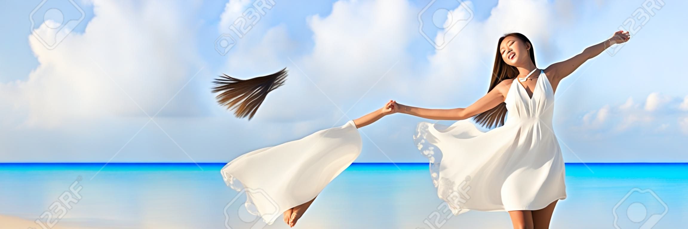 Freedom young woman with arms up outstretched to the sky with blue ocean landscape beach background copy space. Banner panorama. Asian girl in white dress dancing carefree in sunset.