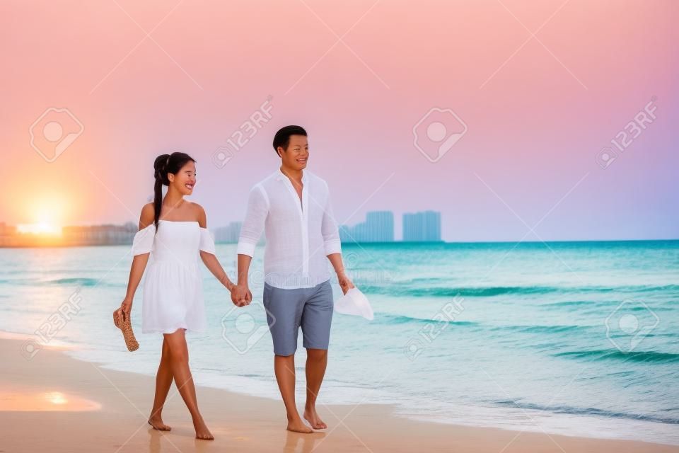 ビーチカップルロマンチックな夕日は、白いドレスとリネンの服を着てフロリダの休暇ビーチ旅行の休日に歩いてリラックスアジアの女性と白人の男を散歩。幸せな異人種間の関係。