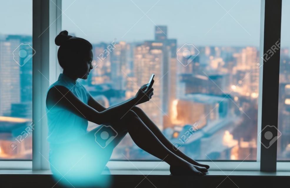 Woman using mobile phone relaxing by condo window at home or office room silhouette. Business woman at work pensive looking at cellphone social media app. Mental health online addiction concept.