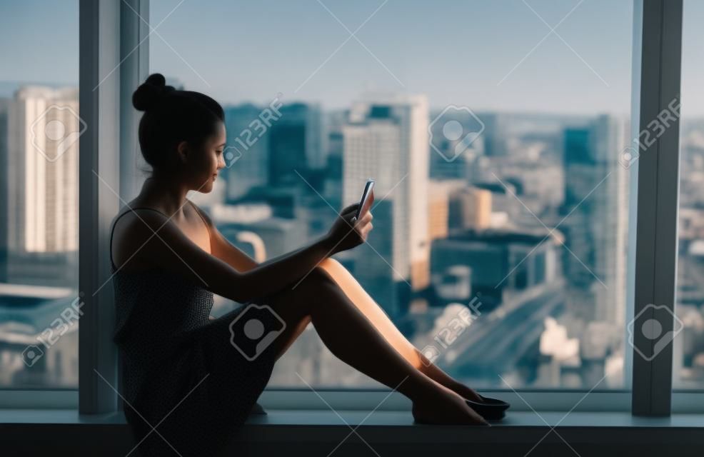 Woman using mobile phone relaxing by condo window at home or office room silhouette. Business woman at work pensive looking at cellphone social media app. Mental health online addiction concept.