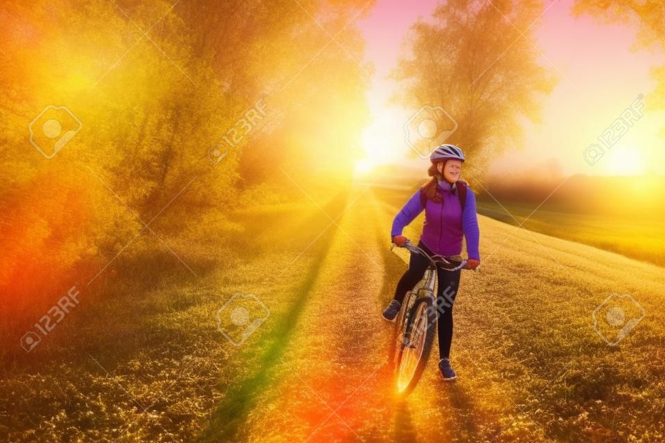 Young bicyclist riding in autumn field at sunset. Happy woman smiling. Active way of life