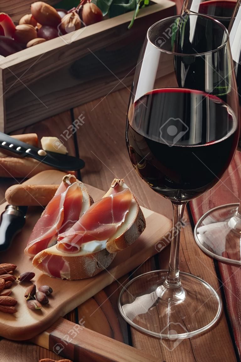 Two glasses of red wine stand on a wooden table. Prosciutto with cheese, red wine, baguette, nuts. Red wine with traditional Mediterranean snacks.