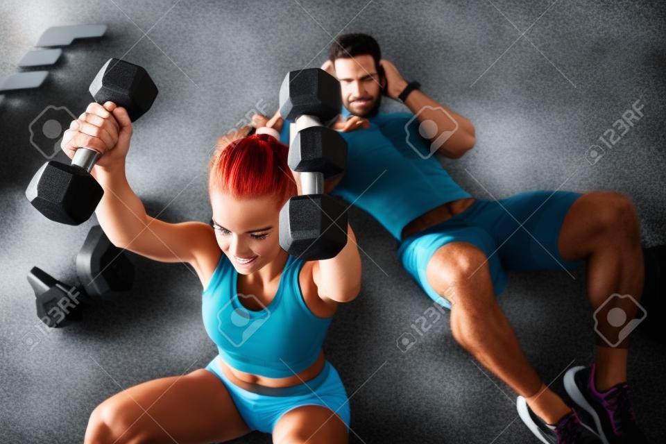 Sportive young couple, man and woman doing abdominal exercises with dumbbells, on the floor in the gym