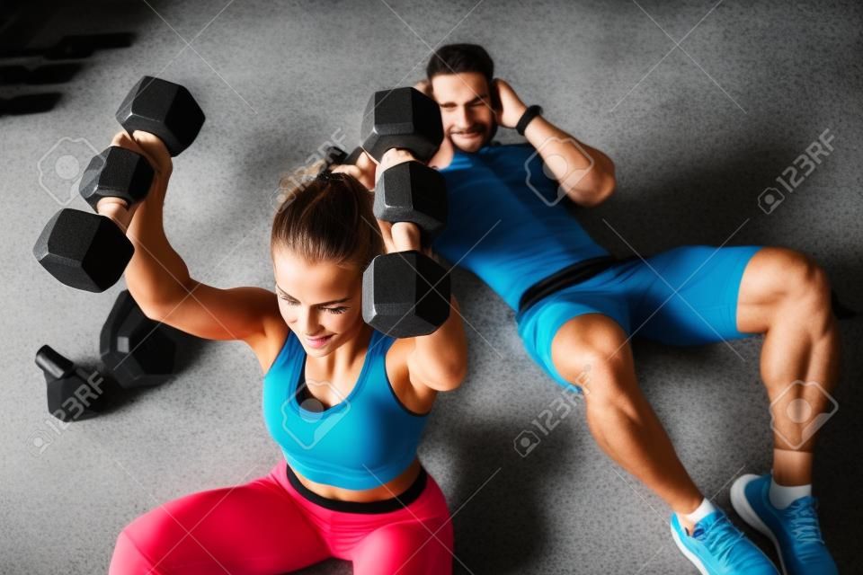 Sportive young couple, man and woman doing abdominal exercises with dumbbells, on the floor in the gym
