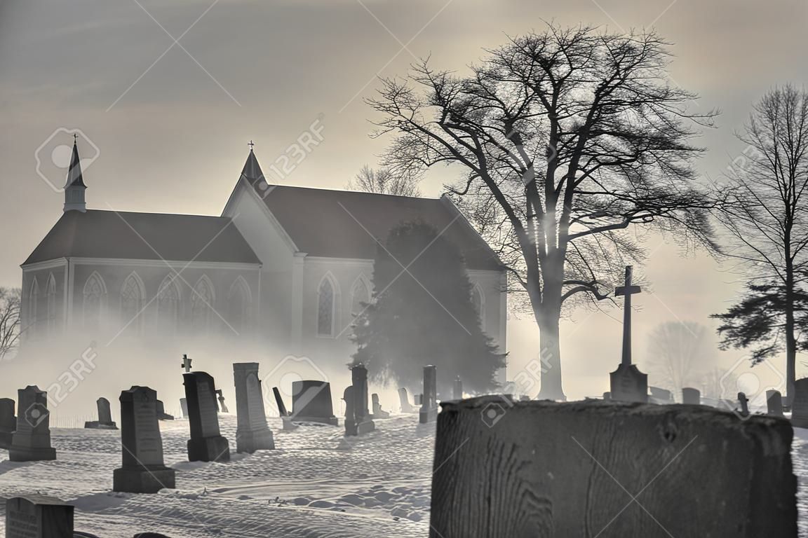 A Cumberland, Maryland church sits behind a graveyard with fog creating an eerie, spooky feeling. Monochromatic image with focus on the gravestones.