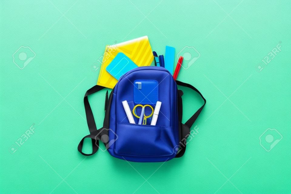 Back to school, education concept. Yellow backpack with school supplies - notebook, pens, ruler, calculator, scissors isolated on blue background. Top view. Copy space Flat lay composition Banner.