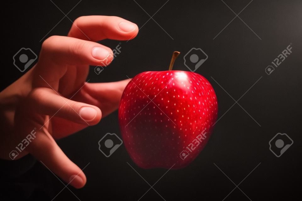 Hand reikend naar een rode appel, de verboden vrucht, in zwarte achtergrond.