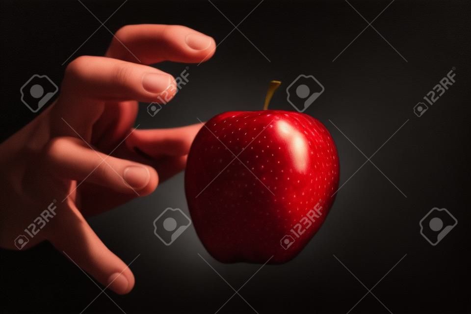 Mão que alcança para uma maçã vermelha, o fruto proibido, no fundo preto.