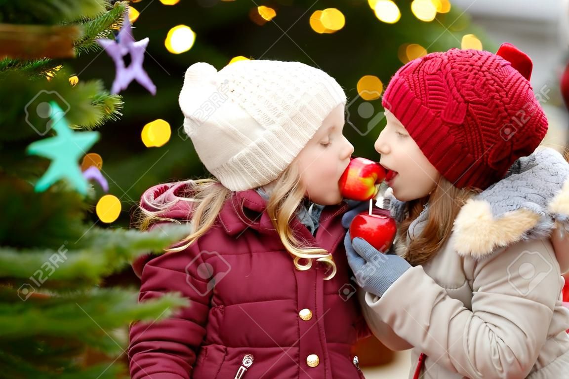 Two adorable little sisters eating red apples covered with sugar icing on traditional Christmas market. Children enjoying sweets, candies and gingerbread on magical Xmas time.