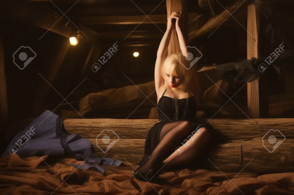 Amarrado a uma menina de madeira do pólo que senta-se em um sótão velho de um edifício abandonado. Menina loira nova bonita em um vestido preto e meias como um refém com suas mãos amarradas