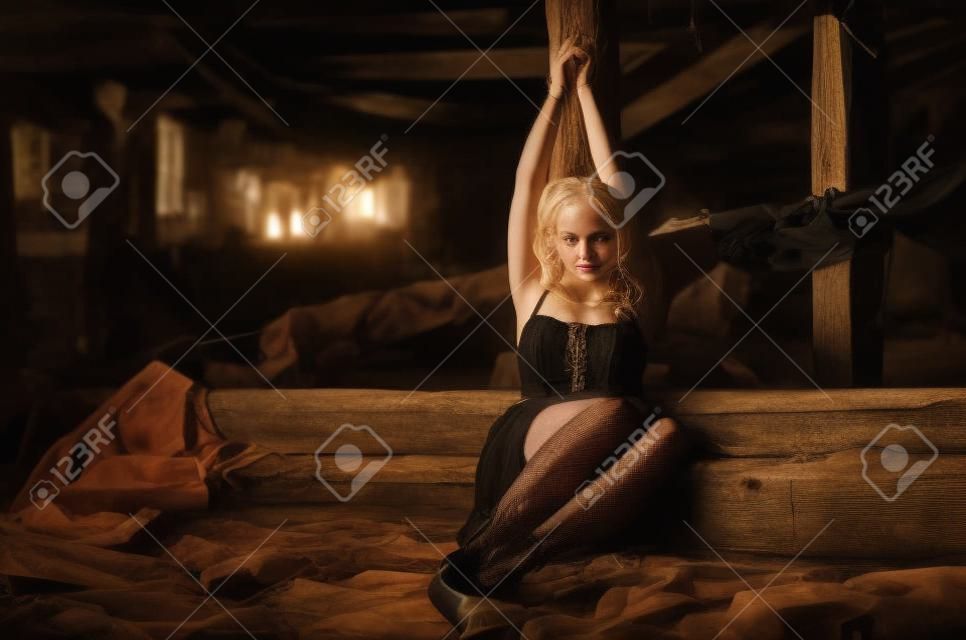 Legata a una ragazza con un palo di legno seduta in una vecchia soffitta di un edificio abbandonato. Bella giovane ragazza bionda in un vestito nero e calze come ostaggio con le mani legate