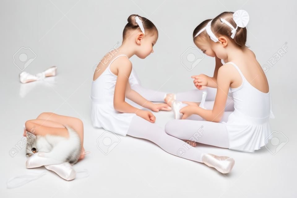 Tres niñas de ballet que se sientan en traje de baño y zapatos blancos de punta junto con el gato en el fondo blanco en el estudio de ballet