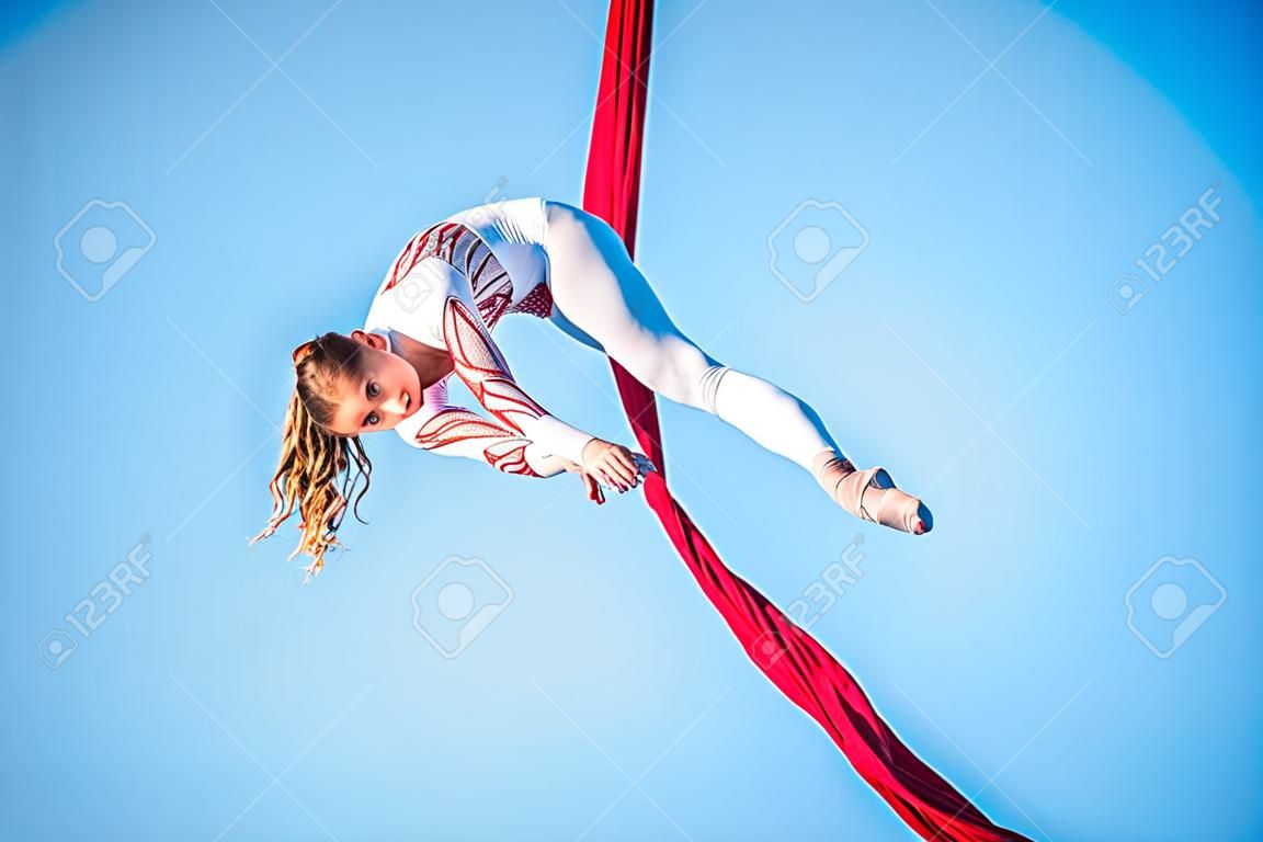 Graceful gymnast performing aerial exercise with red fabrics on blue background